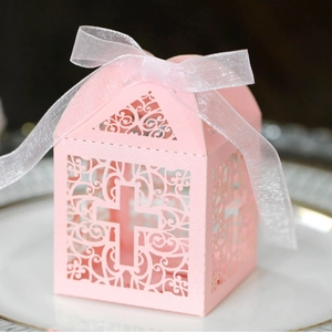 Pink laser-cut candy box with hollow cross design and white ribbon on a white plate