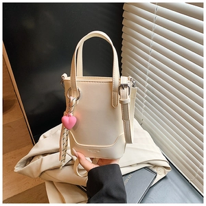 Détail du sac seau crème avec breloque cœur rose tenu contre un trench beige et un livre