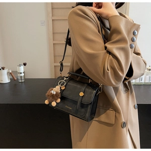 Detail of a black leather crossbody bag featuring a golden logo and a plush doll pendant