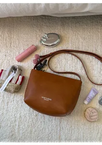 Close-up of caramel leather crossbody bag with cherry charm and brand text on textured background