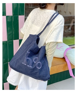 Model wearing a dark blue denim tote bag with white embroidery in a casual street setting