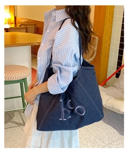 Close-up of a blue denim tote bag with white HO embroidery on a model in a cafe