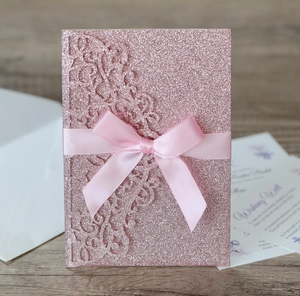 Detailed laser-cut glitter pink invitation with elegant silk ribbon bow on wood background