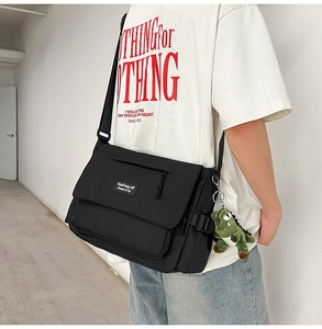 Classic black messenger bag detail with a green dinosaur plush charm on a fabric strap