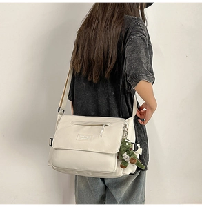 Cream messenger bag side view with green dinosaur plush charm on a wide shoulder strap