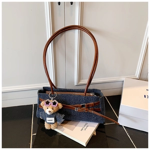 Close-up of blue denim baguette bag with brown leather handles and teddy bear pendant
