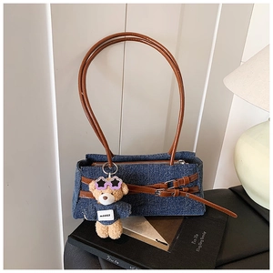 Close-up of denim shoulder bag featuring brown leather straps and a cute teddy bear charm