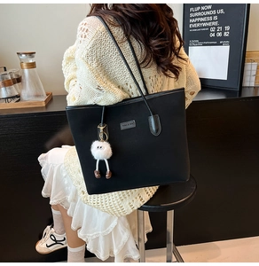 Fashionable black tote bag with white plush doll charm shown in a lifestyle shoulder carry