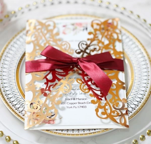 Detail of laser-cut rose gold wedding invitation with a burgundy ribbon on a white plate