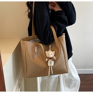 Detail of woven khaki tote bag with plush bear charm and slender shoulder strap