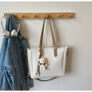 Close-up of white woven tote bag with plush bear charm and brown handles on wooden hook