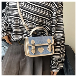 Detail of denim crossbody bag featuring metal heart ornament and leather piping accents