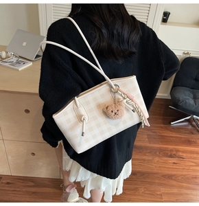 Detail of checkered tote bag with plush bear charm and ribbon on a cream shoulder strap
