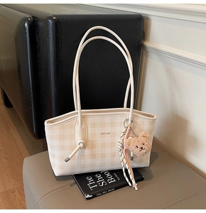 Detail of checkered tote bag featuring dual straps, branding text, and a plush bear charm
