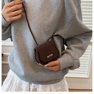 Brown mini PU leather saddle bag with gold 'orin' logo worn over a grey sweatshirt