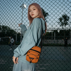 Orange nylon crossbody bag with black strap and bungee detail in outdoor urban setting