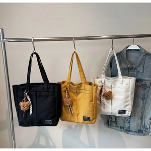 Three minimalist canvas tote bags in black, yellow, and white with front pockets and bear charms
