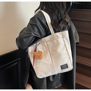 Detail of white canvas tote bag with dual front pockets and brown teddy bear charm