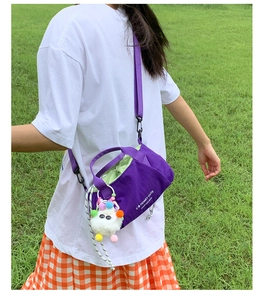 Purple canvas crossbody bag with drawstring closure and plush ball charm on green grass