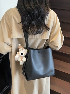 Minimalist black bucket tote bag with plush dog charm shown on a trench coat model