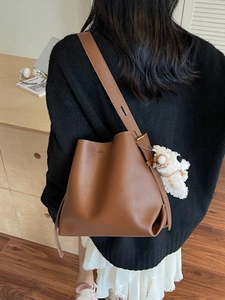 Brown leather bucket bag with wide strap and white plush charm on model in a cozy setting