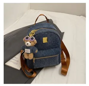 Dettaglio zaino in denim con spallacci marroni e ciondolo a forma di orsetto su letto