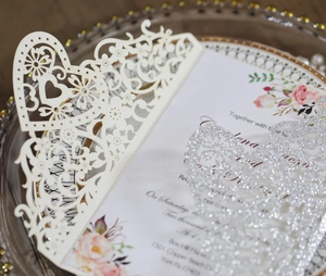Macro view of laser-cut wedding invitation featuring cream floral patterns and silver glitter heart