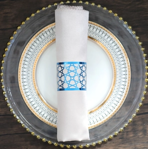 Close-up of blue geometric cutout metal napkin ring on white cloth and gold plate