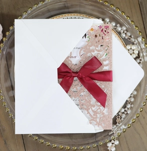 Detail of rose gold glitter laser-cut wedding invitation with burgundy satin bow