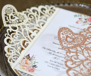 Detail showing laser-cut lace floral pattern and rose gold glitter heart on wedding invitation