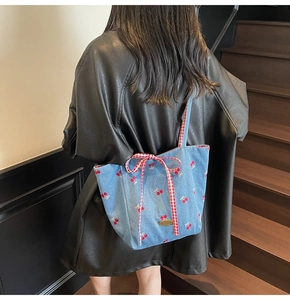 Detail of light blue denim tote bag with red cherry embroidery and a checkered ribbon bow