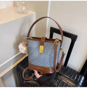 Détail du sac seau en denim avec bordure en cuir marron et pompon blanc sur une valise