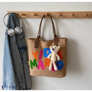Brown canvas tote bag with letter print and plush bear pendant hanging on a hook