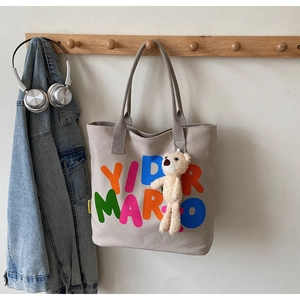 Detail of beige canvas tote bag with YIDR MARIO text and a plush bear charm decoration