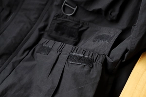 Detail of a tactical black outdoor jacket featuring a large flap pocket with a metal D-ring