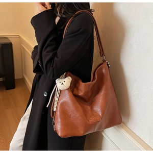 Brown leather tote bag featuring vertical stitching and a cute teddy bear charm accessory