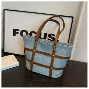 Detailed close-up of large denim tote bag featuring brown leather trim and gold metal rivets