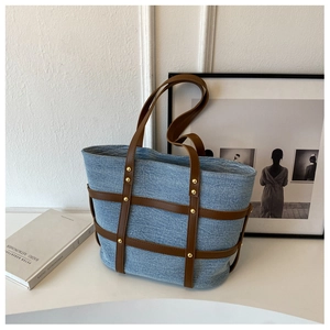Detail of light blue denim tote bag featuring brown leather straps and gold metal rivets