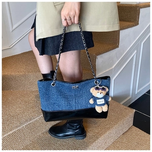 Detail of denim tote bag with leather trim, metal chain strap, and plush bear charm