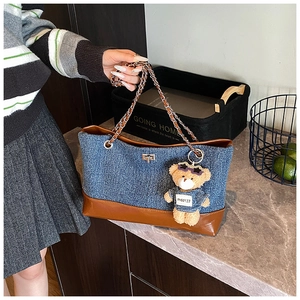 Detail of denim tote bag featuring brown leather base and teddy bear charm accessory