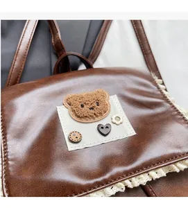 Detail of a brown leather backpack with lace trim, plush bear patch, and heart button