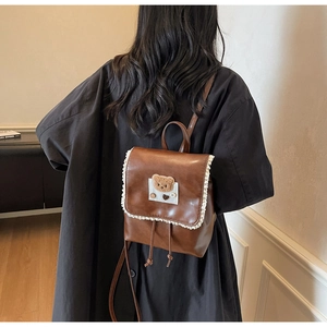 Detail of a brown leather backpack with lace trim and bear patch worn by a model