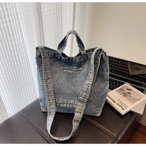 Blue denim tote bag with white text details on a dark leather bench with a laptop