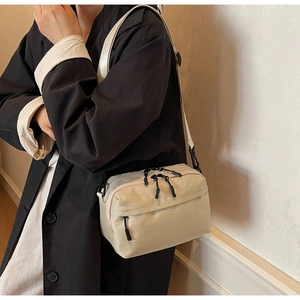 Detail of beige nylon crossbody bag with dual zippers worn over a black trench coat