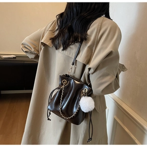 Détail du sac seau marron foncé avec chaîne dorée et boule en peluche sur un trench-coat