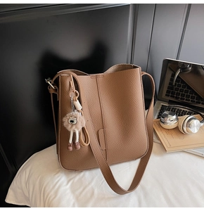 Brown lychee grain bucket bag with a cute plush cartoon charm on white bedding