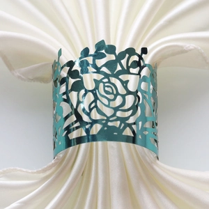 Close-up of teal laser-cut metal napkin ring with rose pattern on white satin