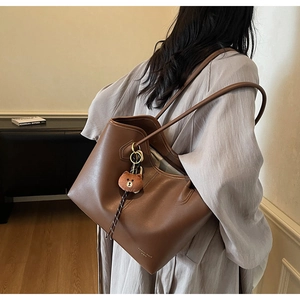 Detail of a large brown leather tote bag with a bear charm worn by a model in a room