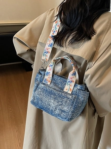 Blue denim tote bag with floral embroidered strap shown on a woman in a trench coat