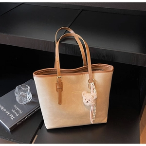 Detail view of large beige tote bag with brown straps and plush bear charm on a black shelf